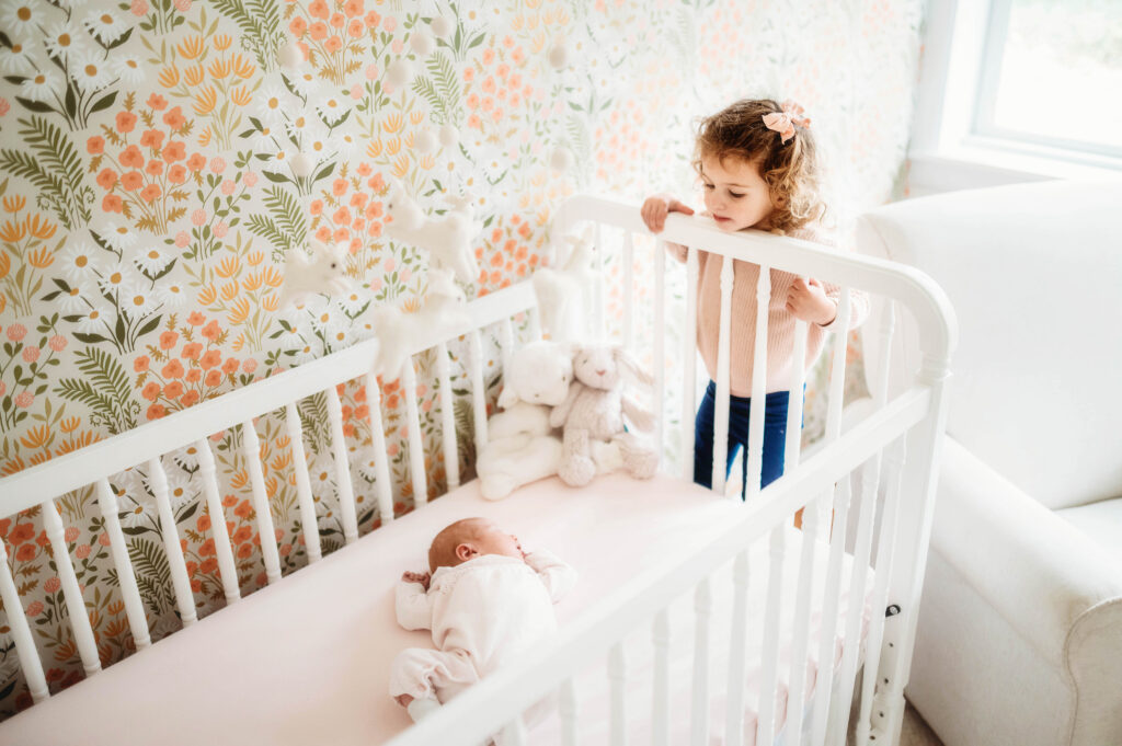 After deciding betweenCharleston Studio Newborn Photography vs. In-Home Lifestyle Sessions, Older sibling peers at her infant baby sister sleeping in her crib, in their home during an in-home Lifestyle Newborn Portrait Session in Charleston, SC.