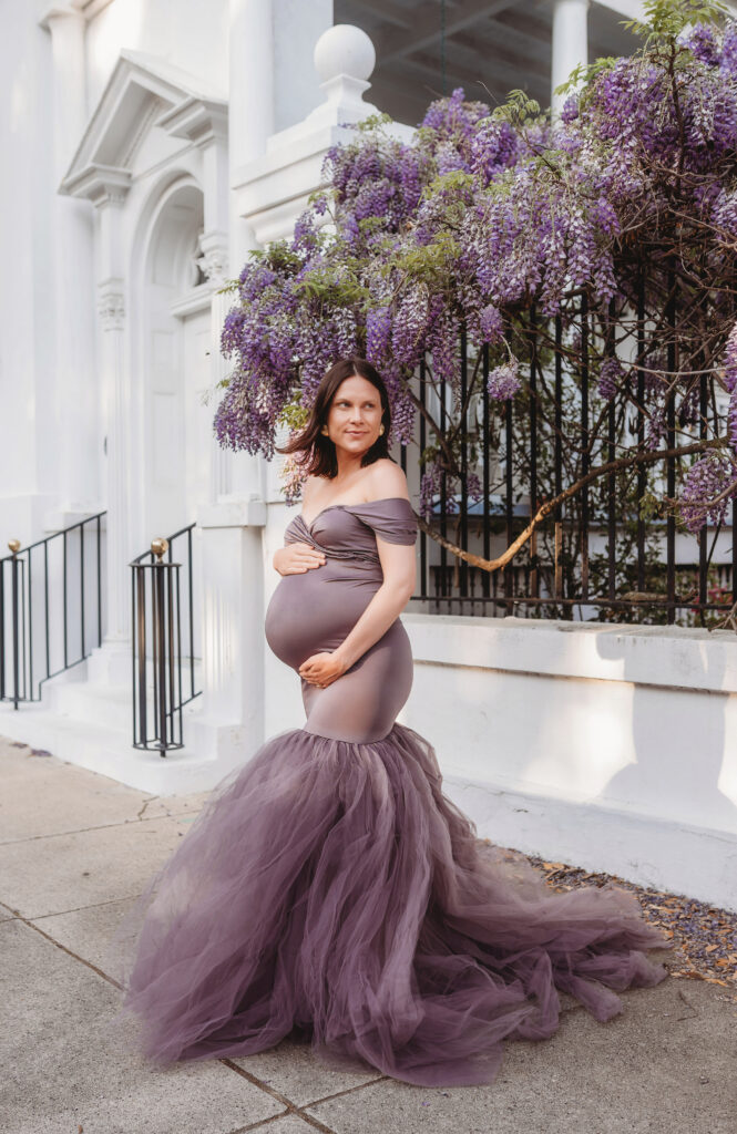 Expectant mother poses for Spring Maternity Portraits beneath beautiful purple wisteria blooms in downtown Charleston, SC.
