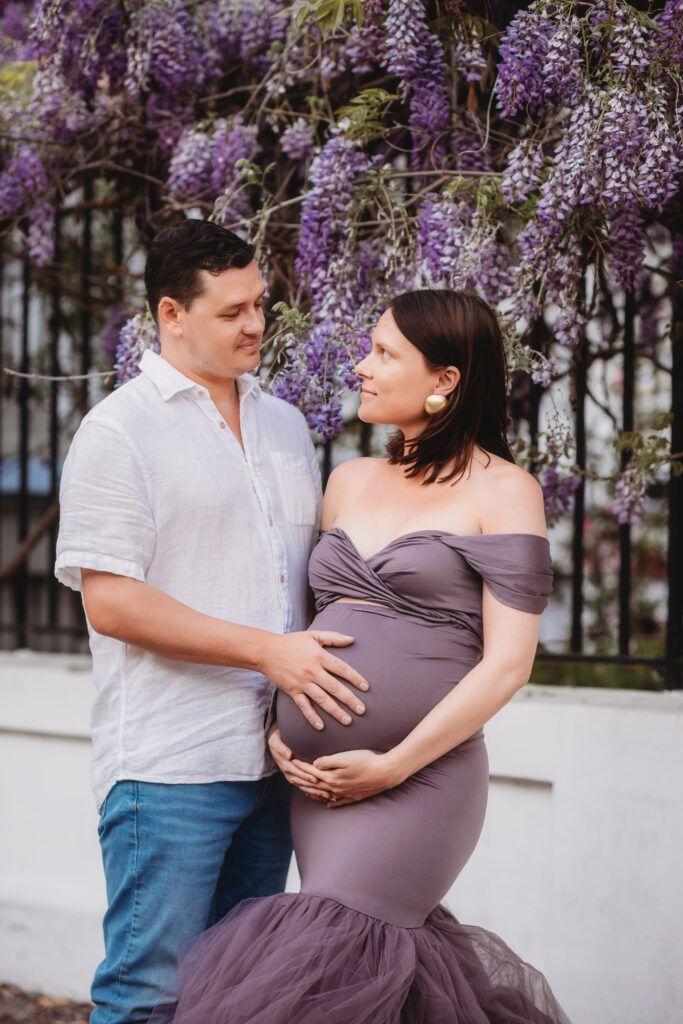 Expectant parents pose for Spring Maternity Portraits beneath beautiful purple wisteria blooms in downtown Charleston, SC.