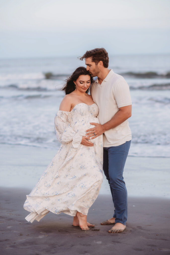 Expectant parents share a kiss together on Folly Beach during their Maternity Portrait Session on Folly Beach after discovering The Ultimate Charleston Babymoon Planning Guide.
