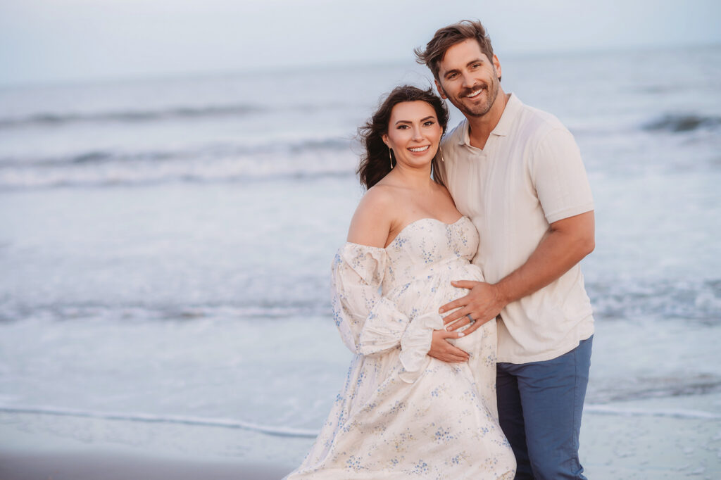 Expectant parents pose together on Folly Beach during their Maternity Portrait Session on Folly Beach after discovering The Ultimate Charleston Babymoon Planning Guide.