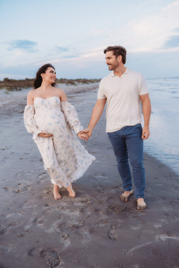 Expectant parents walk together on Folly Beach during their Maternity Portrait Session on Folly Beach after discovering The Ultimate Charleston Babymoon Planning Guide.