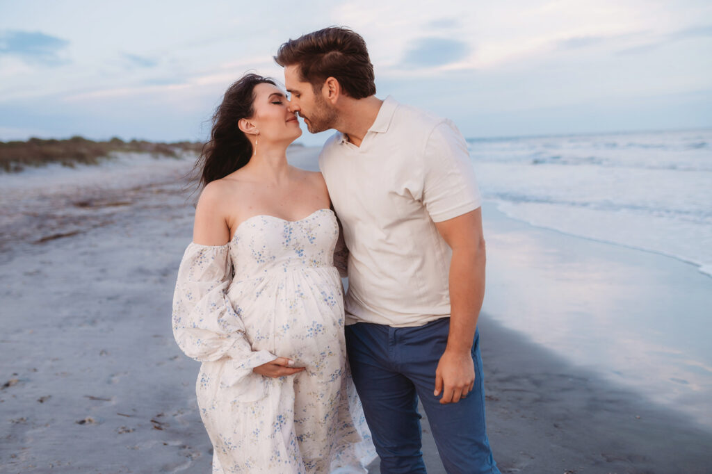 Expectant parents pose together on Folly Beach during their Maternity Portrait Session on Folly Beach after discovering The Ultimate Charleston Babymoon Planning Guide.